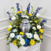 White roses, blue flowers, and yellow daisies arranged around a silver urn