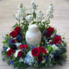 Red roses and white flowers arranged around a white urn on a wooden surface.