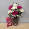 Pink and white flowers in a polka-dot vase with a chocolate box beside it.