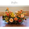 Low floral centerpiece with orange, yellow, and peach blooms in a shallow container