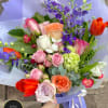 Hand-tied bouquet of mixed roses, tulips, and purple delphinium wrapped in lilac paper.