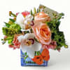 Short peach and white flower arrangement with roses and orchids in a blue glass cube vase with an orange butterfly.