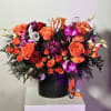 Orange and purple floral arrangement with roses, orchids, and Halloween skeleton hand accents in a black container