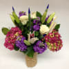 Bouquet of white roses, purple blooms, and lily buds in a burlap-wrapped vase