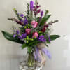 Tall arrangement of pink roses and purple flowers in a clear glass vase with decorative bow