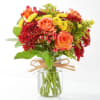 Compact bouquet of orange roses, red mums, and yellow blooms in a clear glass jar with raffia bow.