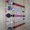 Five decorated pens with beads and charms arranged on a wooden surface.