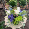 Low arrangement of white roses, purple mums, blue blooms, and green hydrangea in a vase on a small table