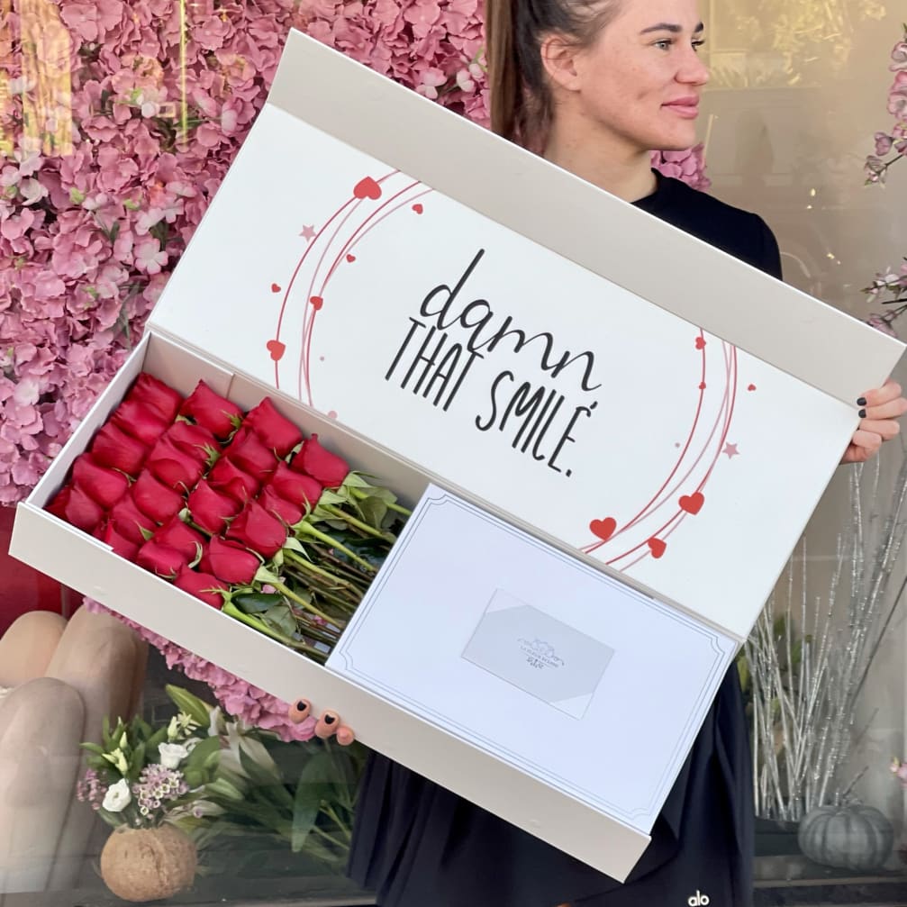 Woman holding a large gift box of red roses with a "damn that smile" message.