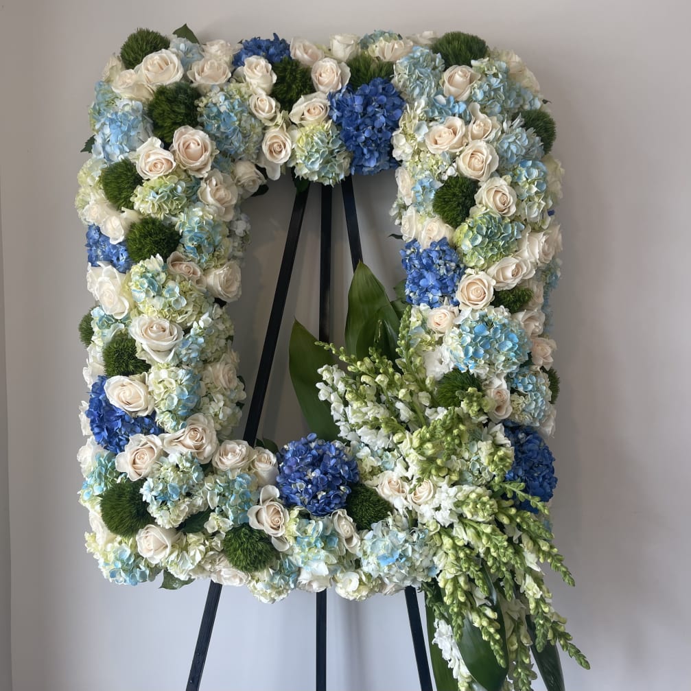 Square floral wreath on an easel with white roses, blue hydrangeas, and green accents