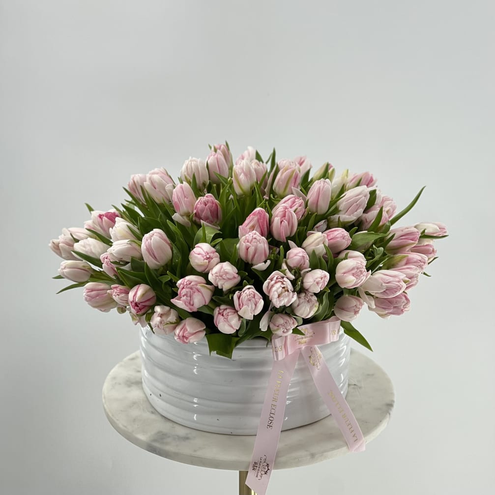 Pink tulips arranged in a white round container with a pink ribbon