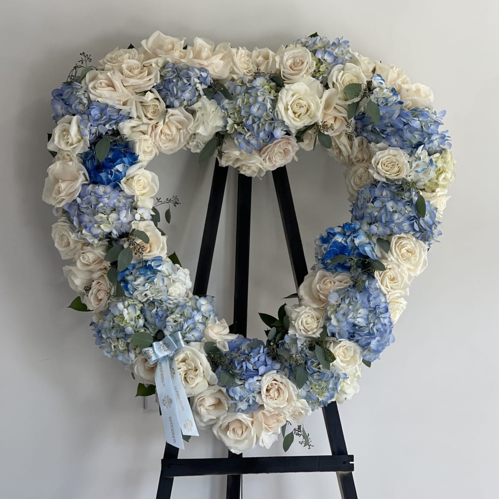 Heart-shaped floral wreath with white roses and blue hydrangeas on a black stand