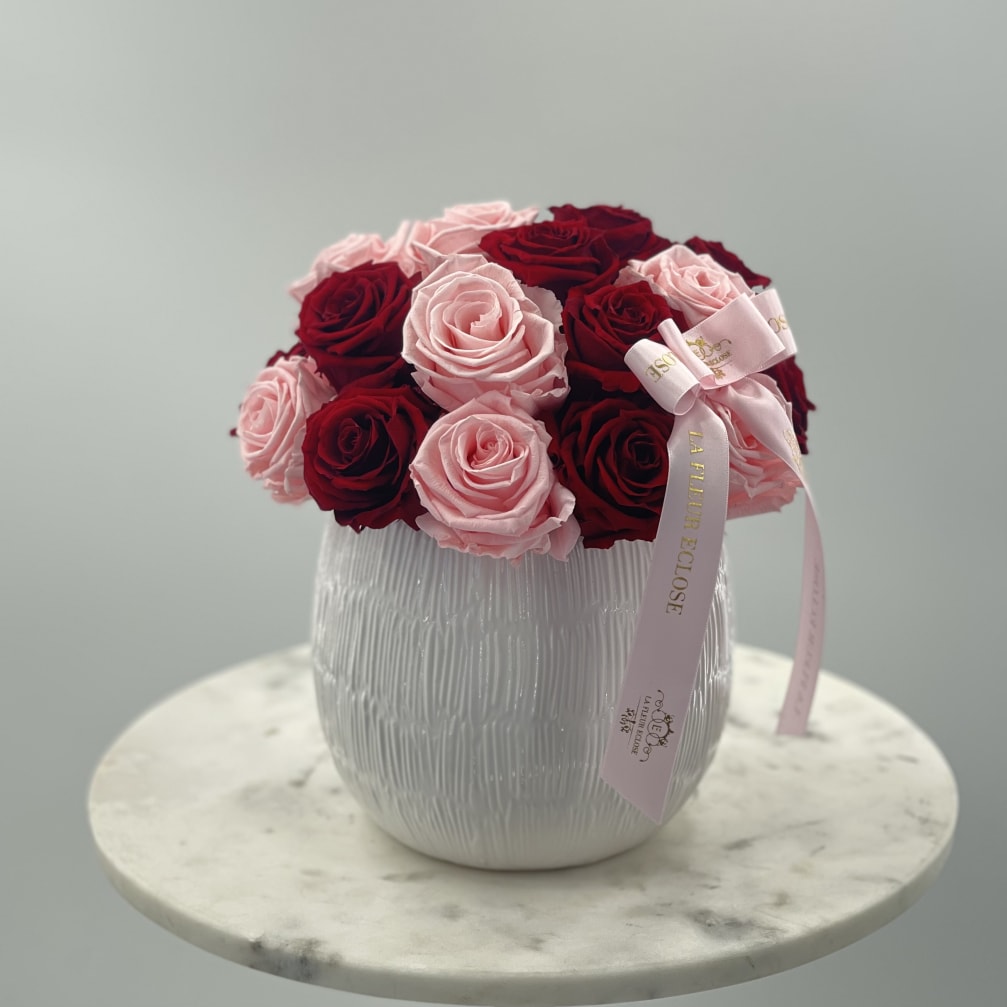 Pink and red roses arranged in a white vase with a pink ribbon