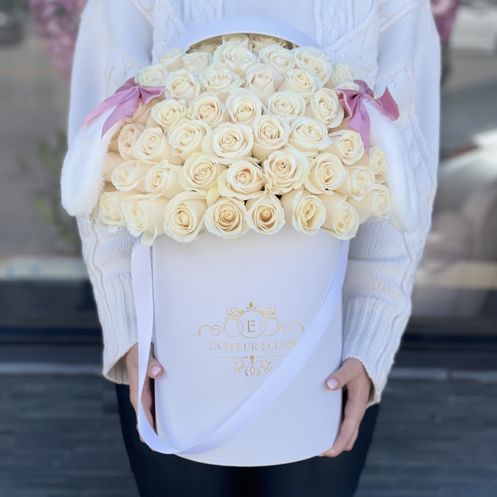Large hat box of cream roses with pink ribbon accents held by a person in a white sweater
