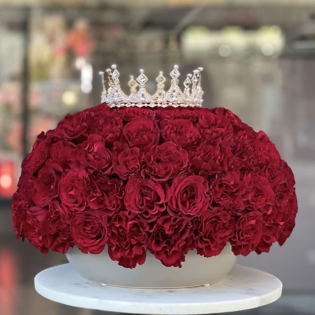 Red rose arrangement topped with a jeweled crown in a white container