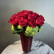 Red roses arranged in a tall red glass vase with green accents