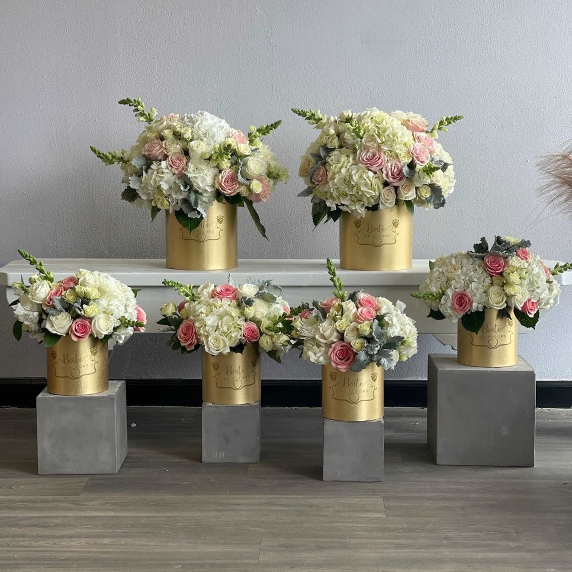 Five pastel floral arrangements in gold cylindrical boxes on gray pedestals