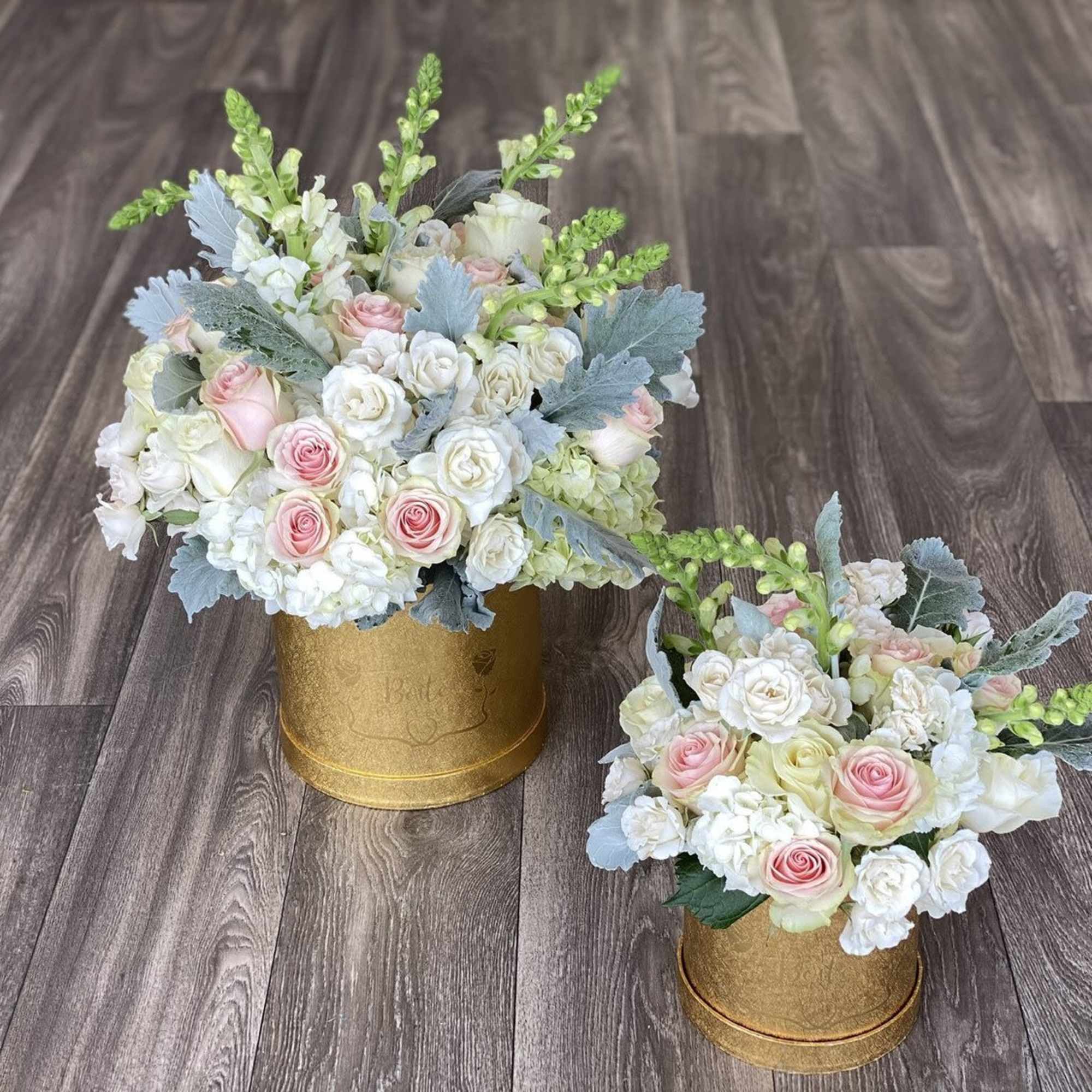 Two pastel rose arrangements in gold containers on a wood floor