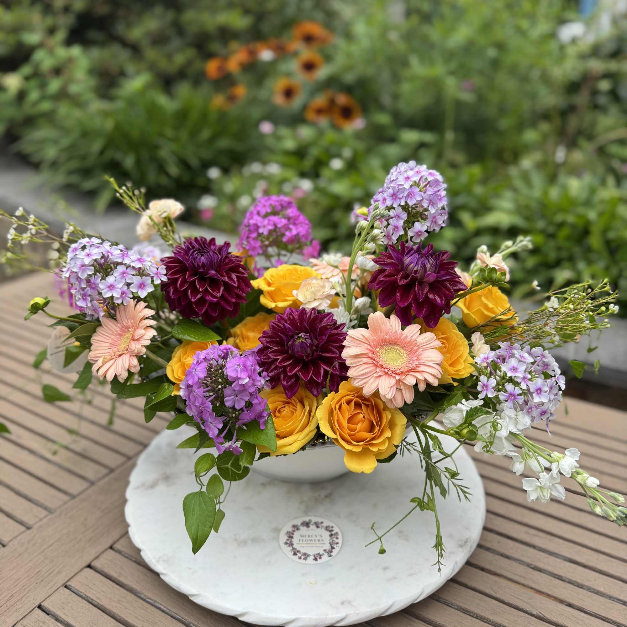 Mixed bouquet of yellow roses, purple blooms, and peach daisies in a white bowl