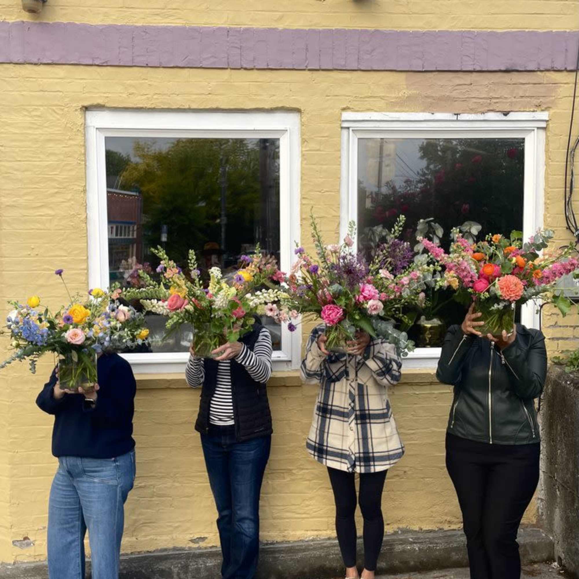 Four people hold large colorful flower arrangements in front of their faces outside a flower shop.