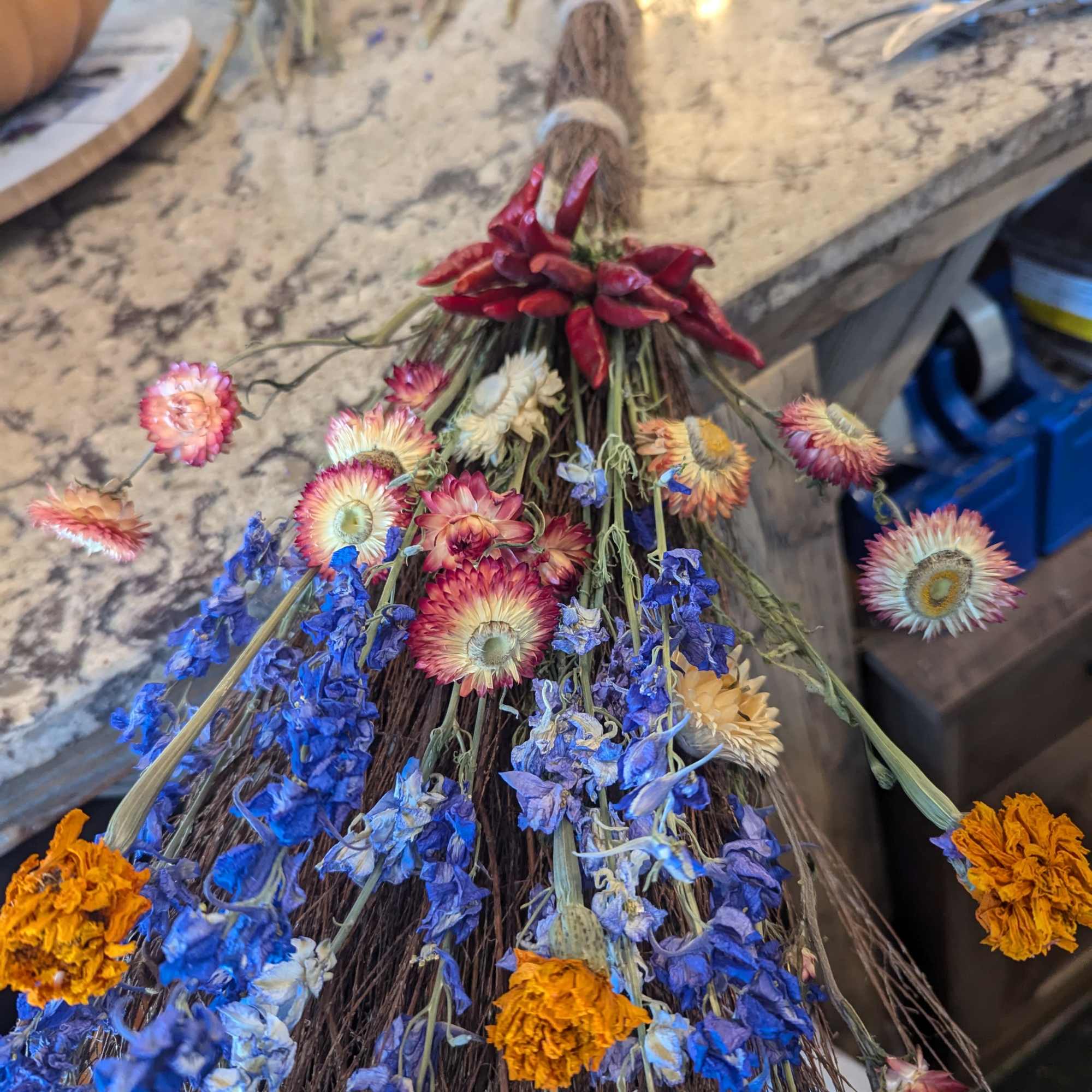 Rustic twig broom decorated with dried blue, orange, and cream flowers and red peppers.