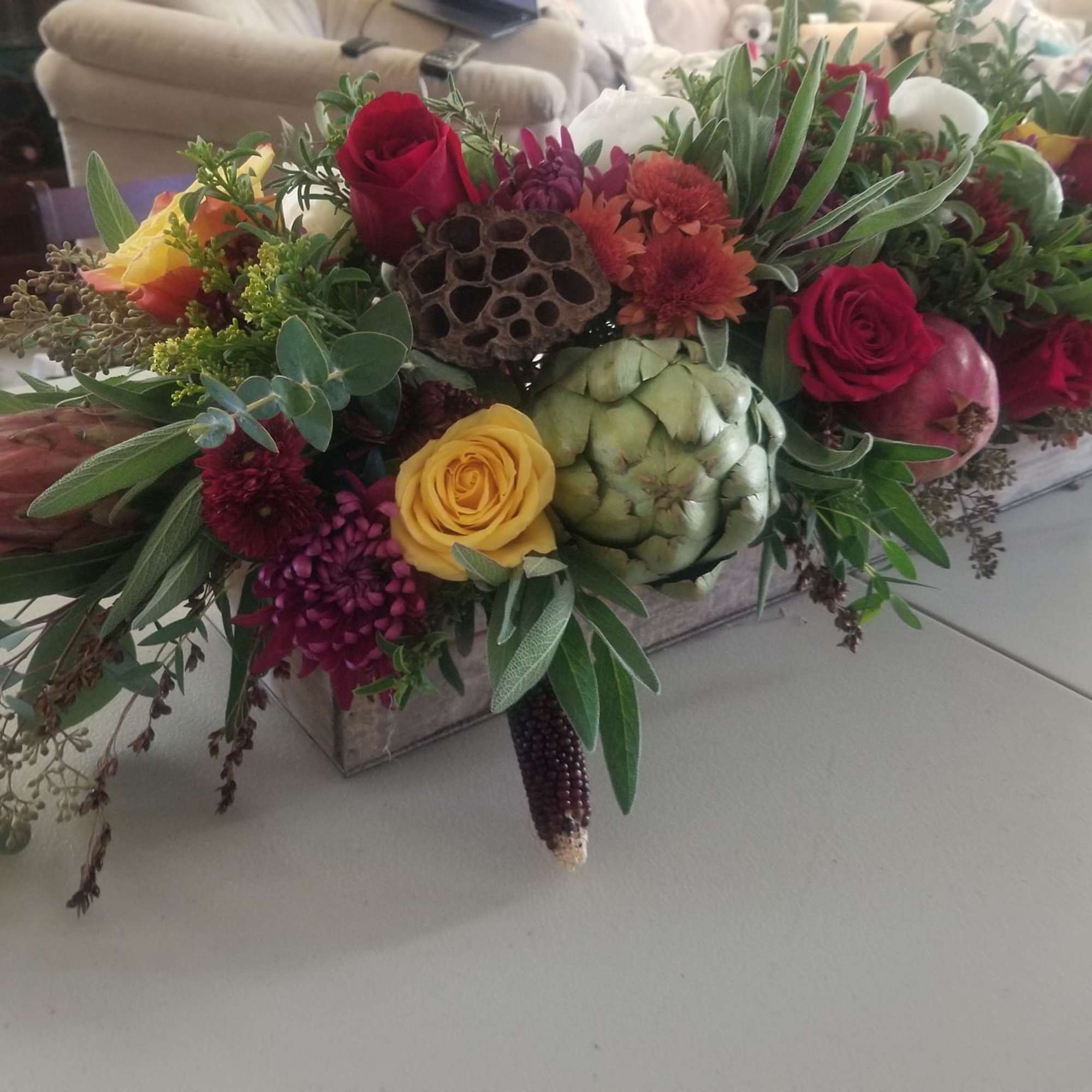 Low centerpiece of red and yellow roses, mums, protea, herbs, and artichoke in a rustic rectangular box