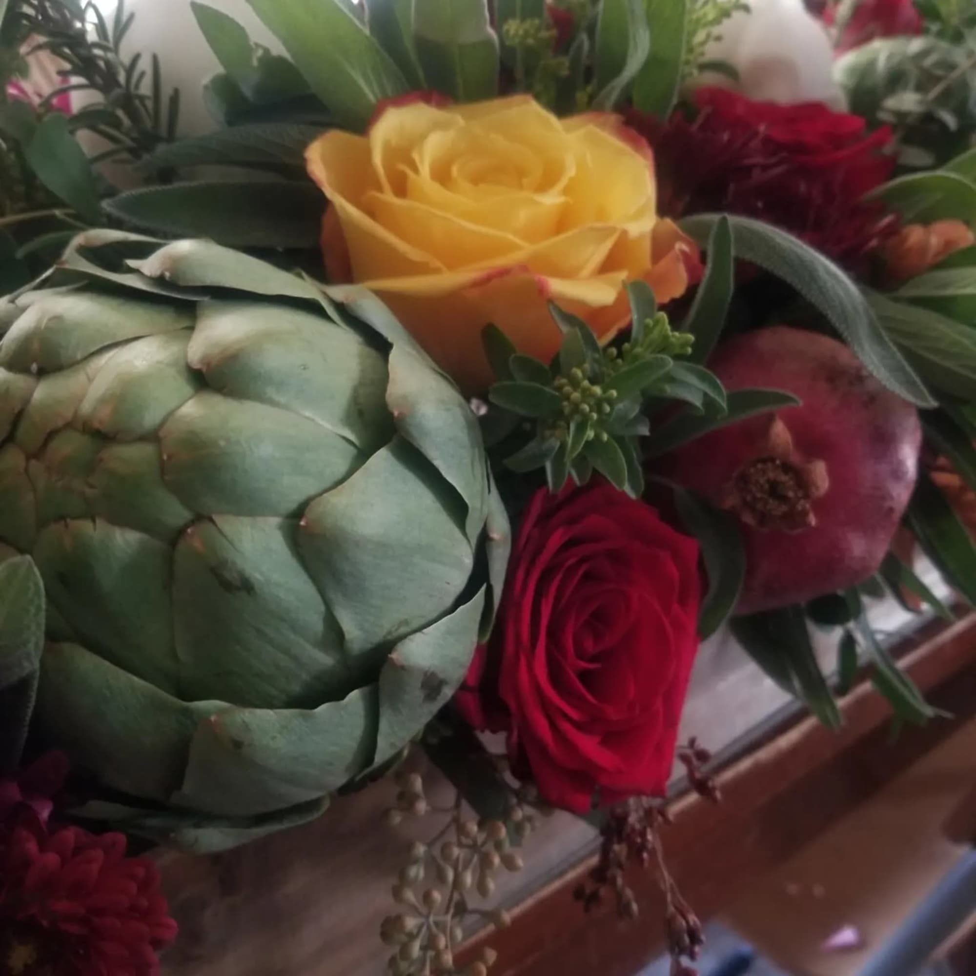 Close-up of arrangement with yellow and red roses alongside an artichoke and a pomegranate.