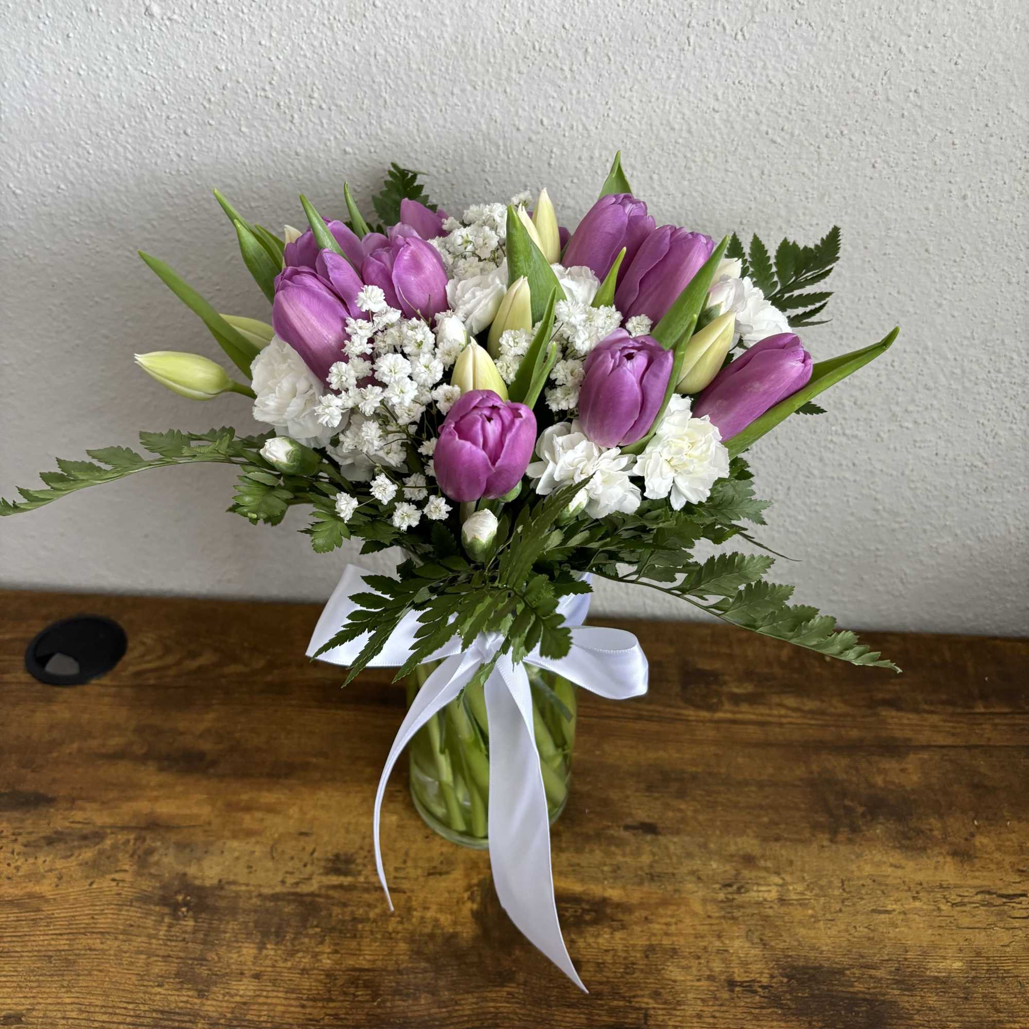 Purple tulips and white flowers in a glass vase with a white ribbon