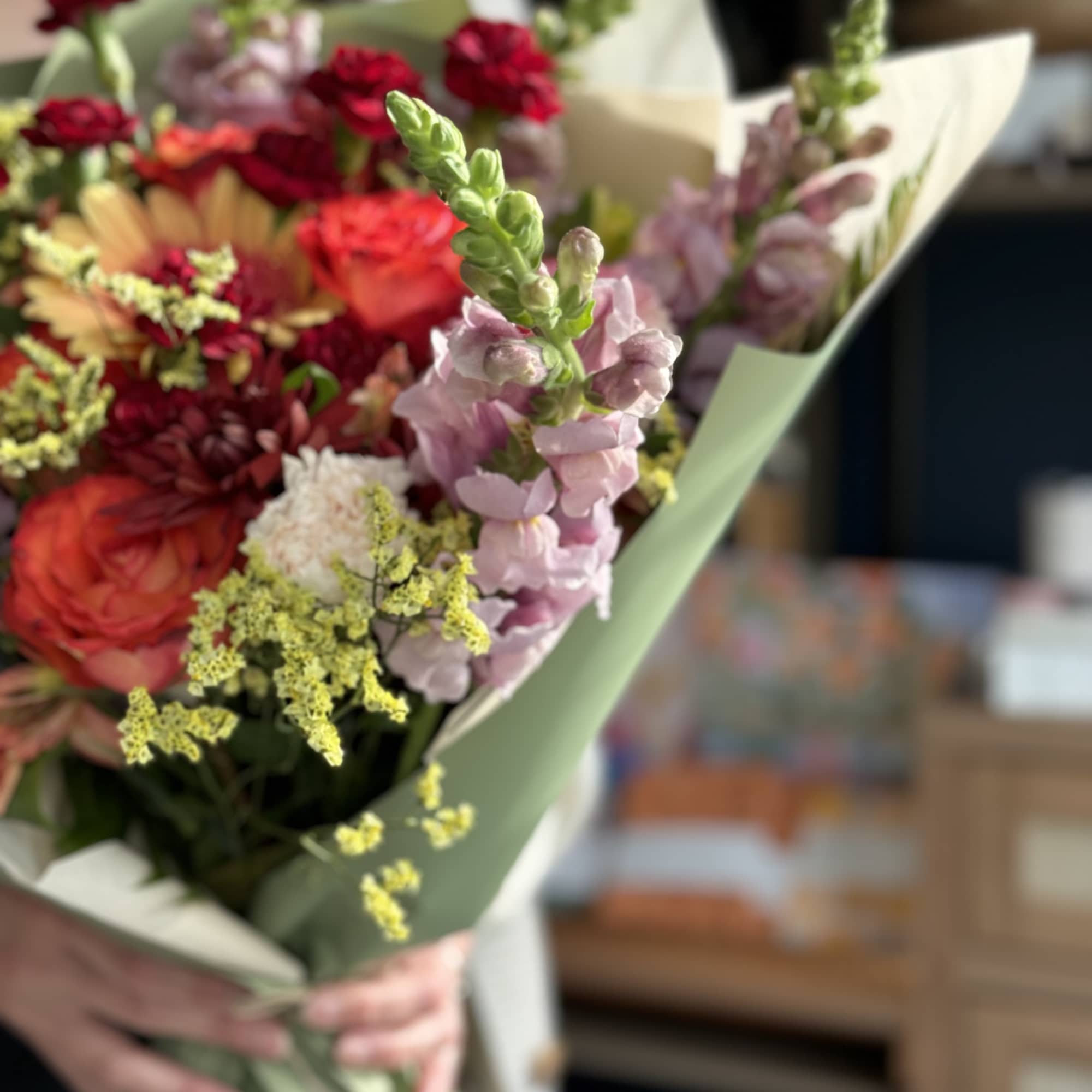 Handheld bouquet of mixed pink, red, and peach flowers wrapped in pale green paper