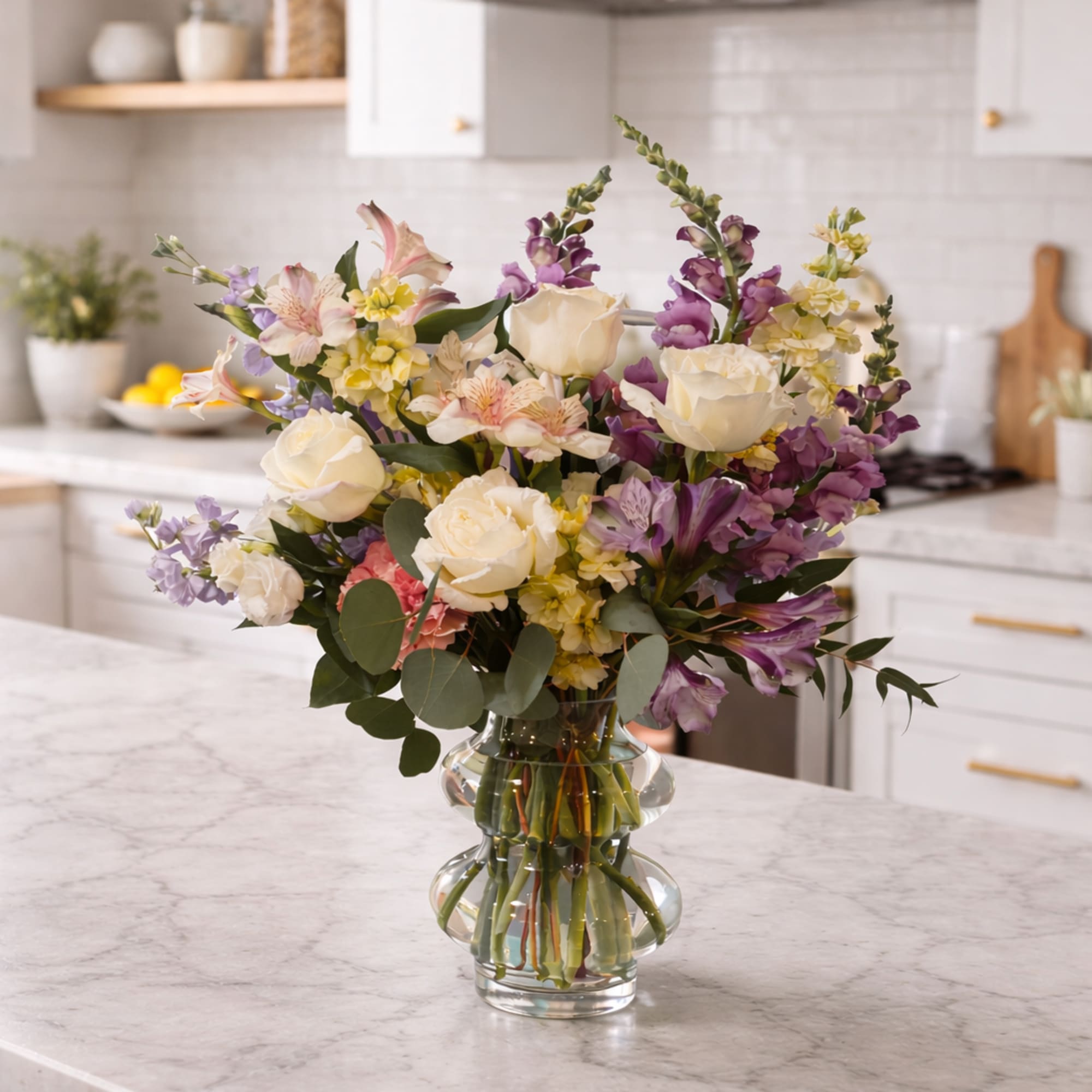 Mixed bouquet of white roses, purple snapdragons, and pastel blooms in a clear glass vase on a kitchen counter