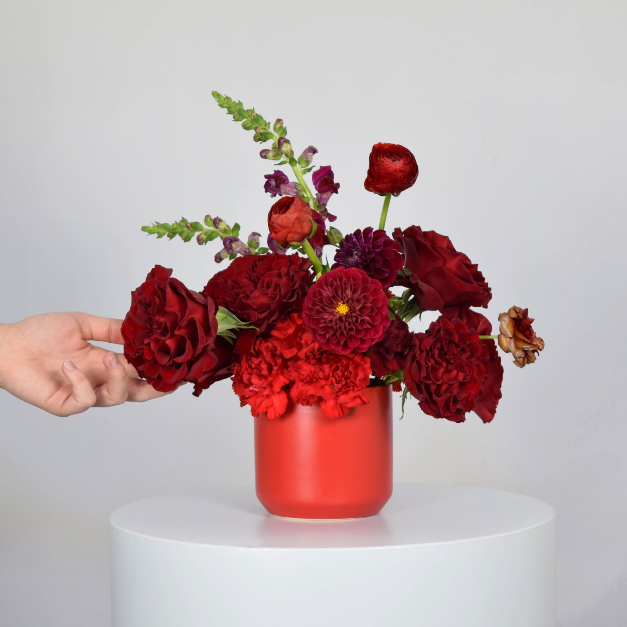 Red floral arrangement in a red vase with a hand beside it