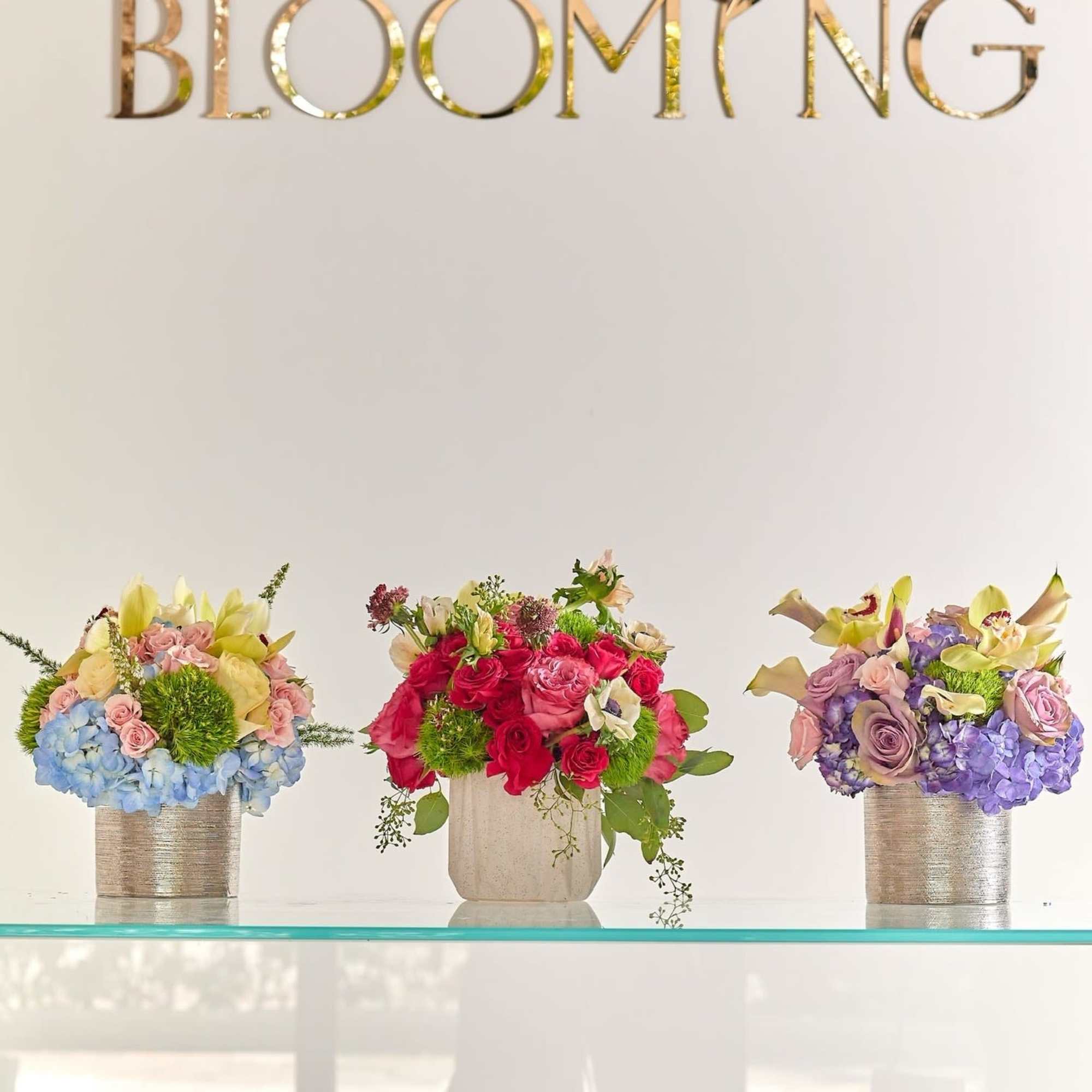 Three colorful floral arrangements in metallic vases beneath a "BLOOMING" sign