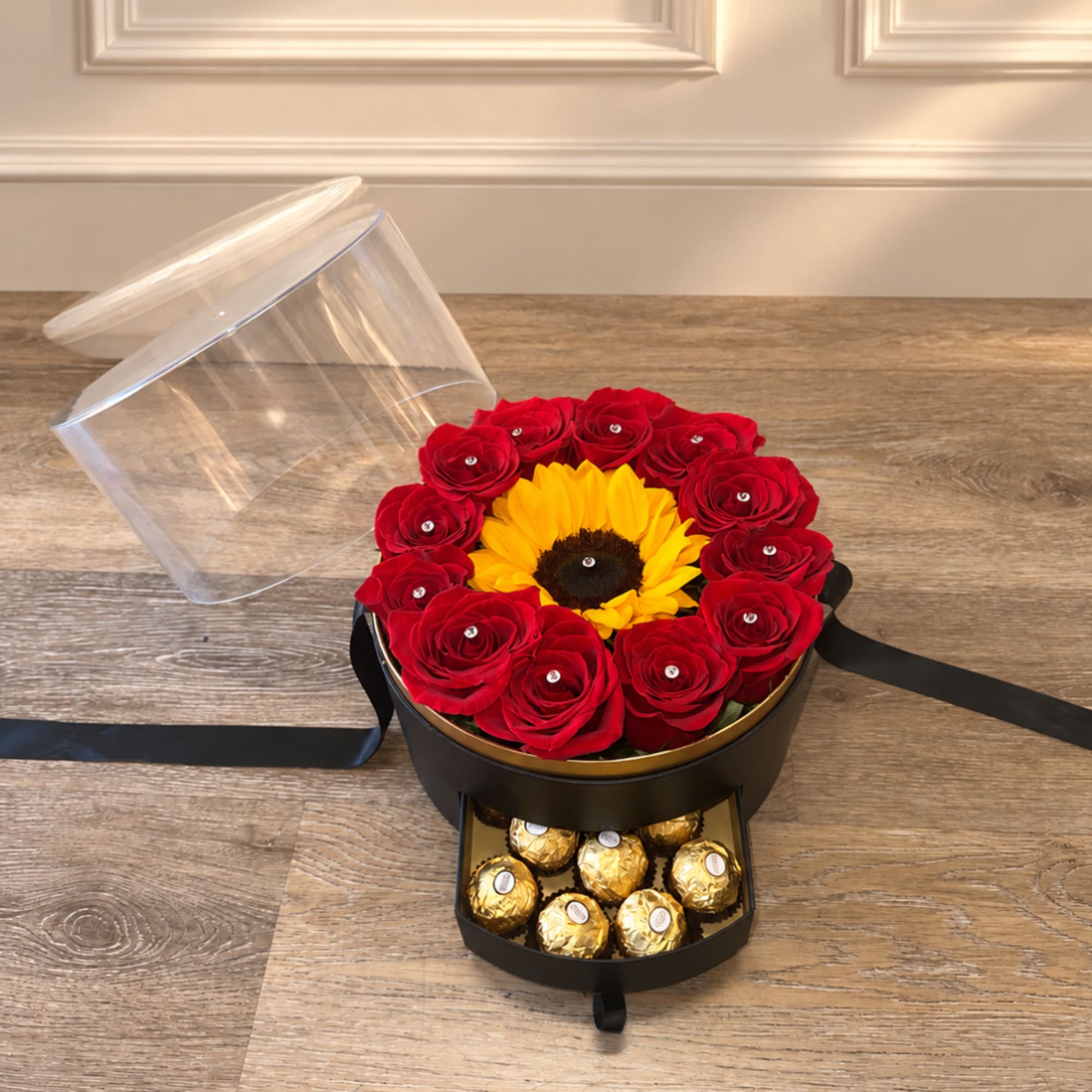 Red roses arranged around a sunflower in a round gift box with chocolates below.