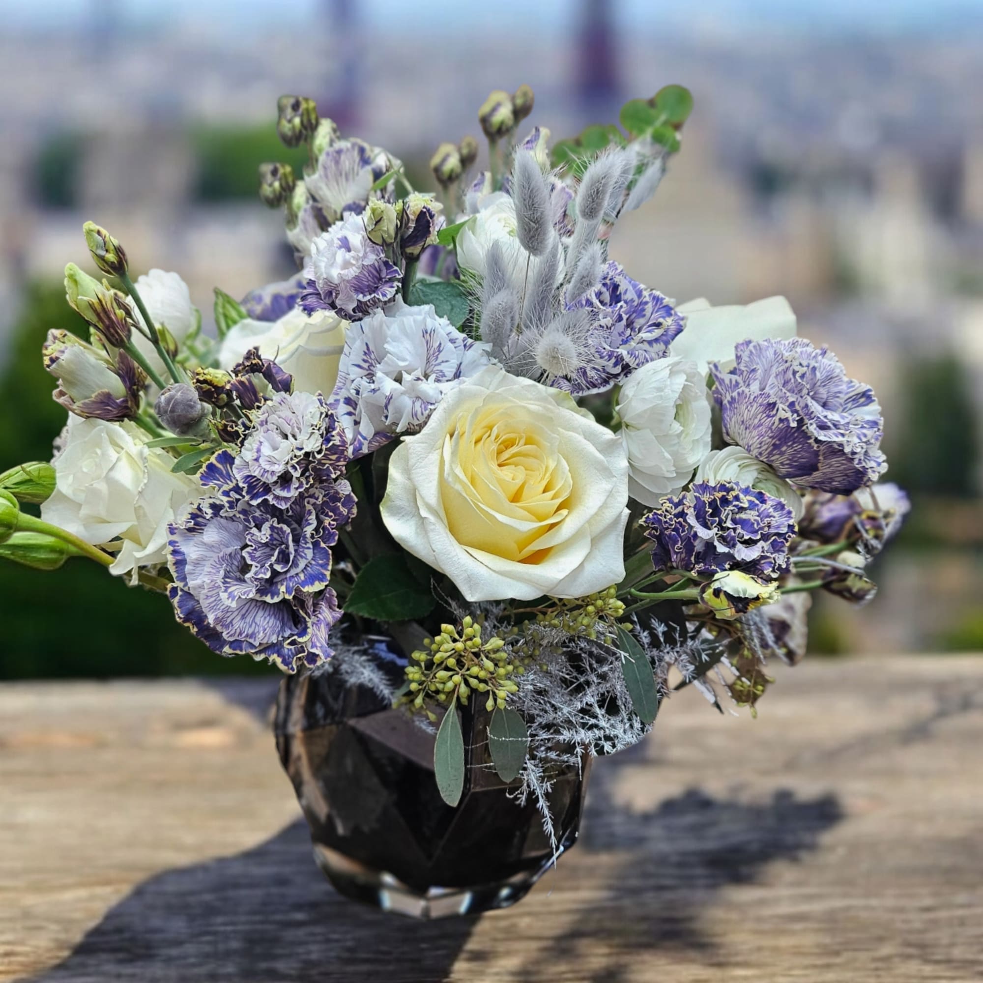 Compact arrangement of cream roses and purple ruffled blooms in a dark faceted glass vase