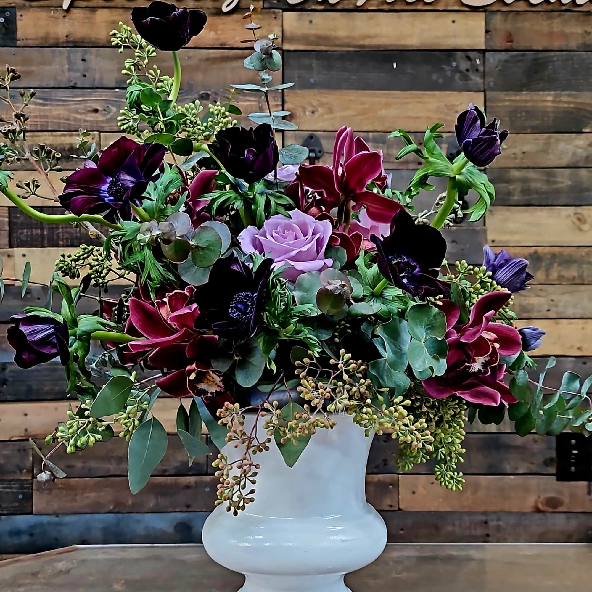 Mixed floral arrangement in a white pedestal vase with dark blooms and lavender roses