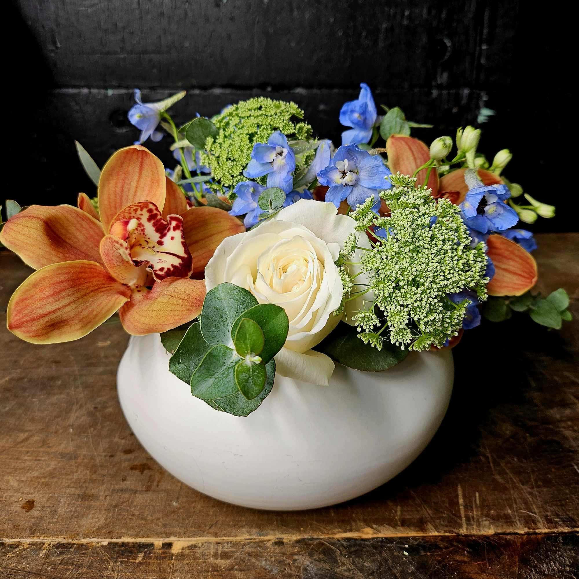 Mixed floral arrangement in a white bowl vase with orange orchids, white rose, and blue flowers