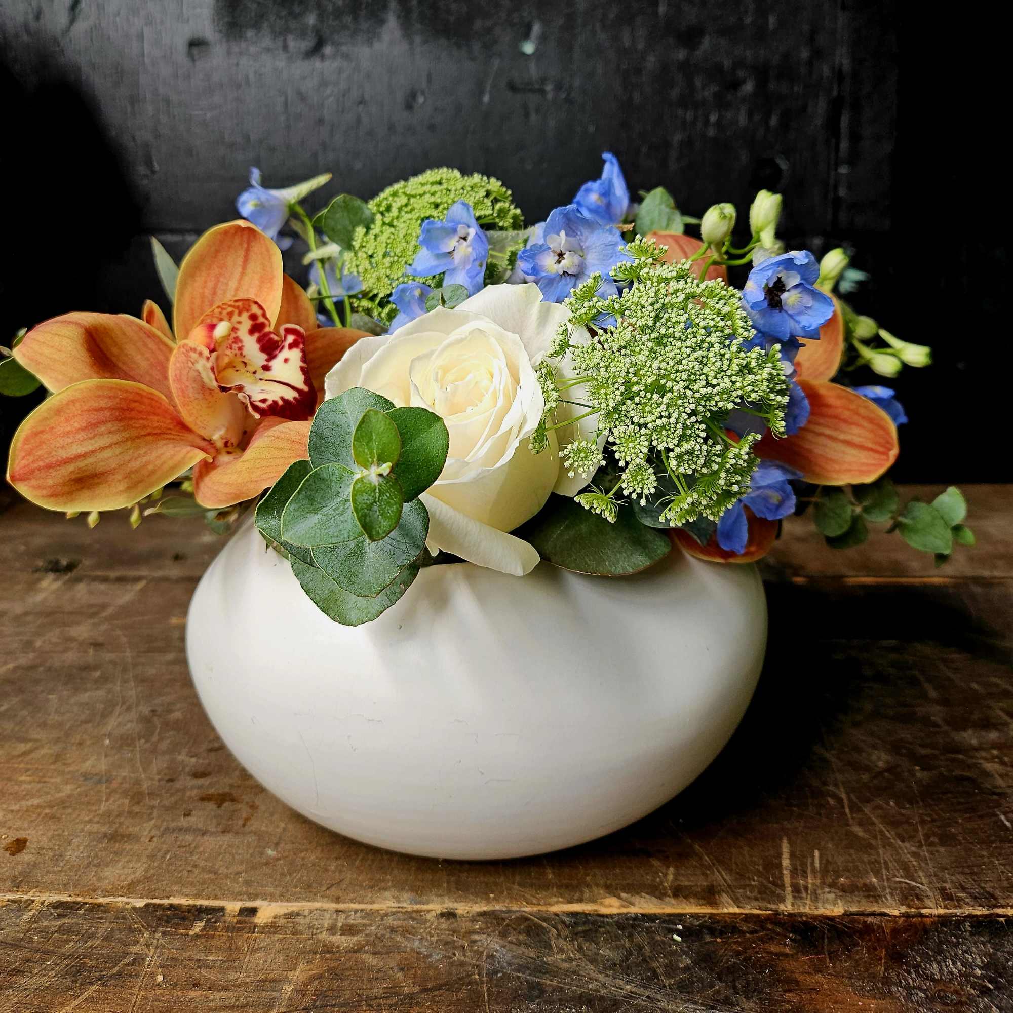 Bouquet of orange orchids, blue flowers, and a white rose in a white vase