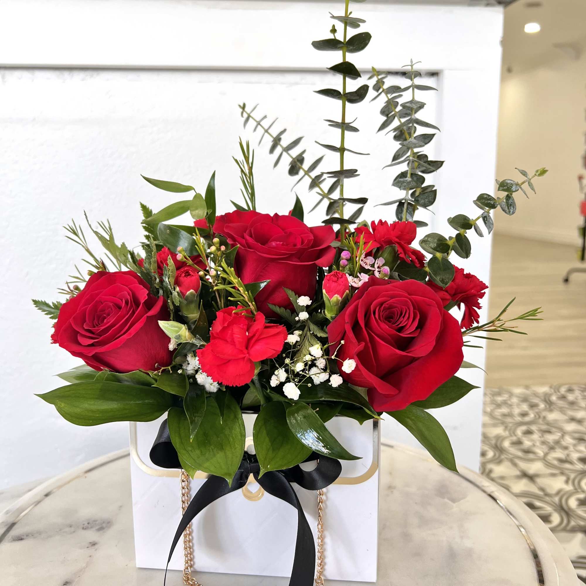 Red roses arranged in a white gift box with greenery and a black ribbon