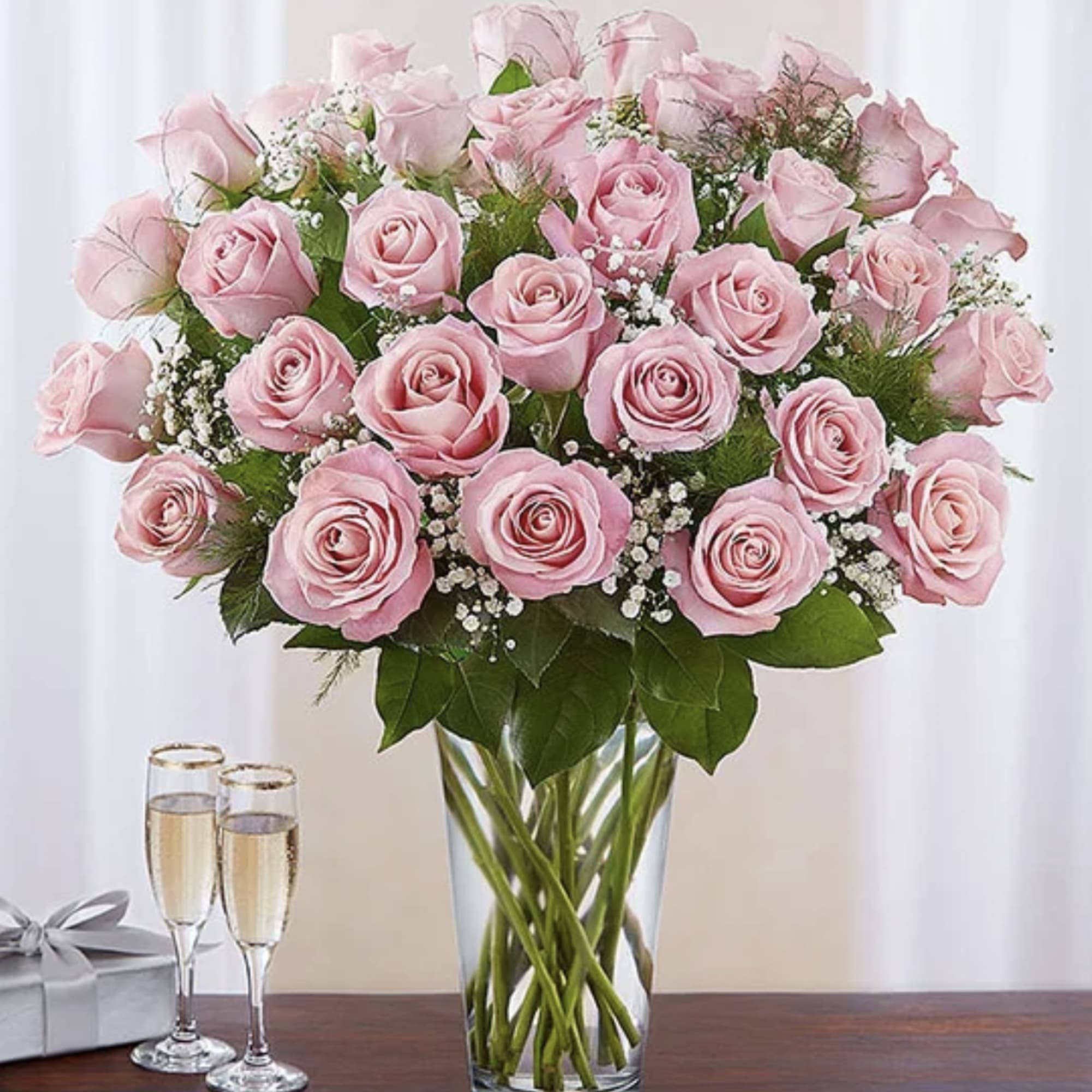 Tall arrangement of light pink roses with baby's breath in a clear glass vase beside champagne glasses and a gift box