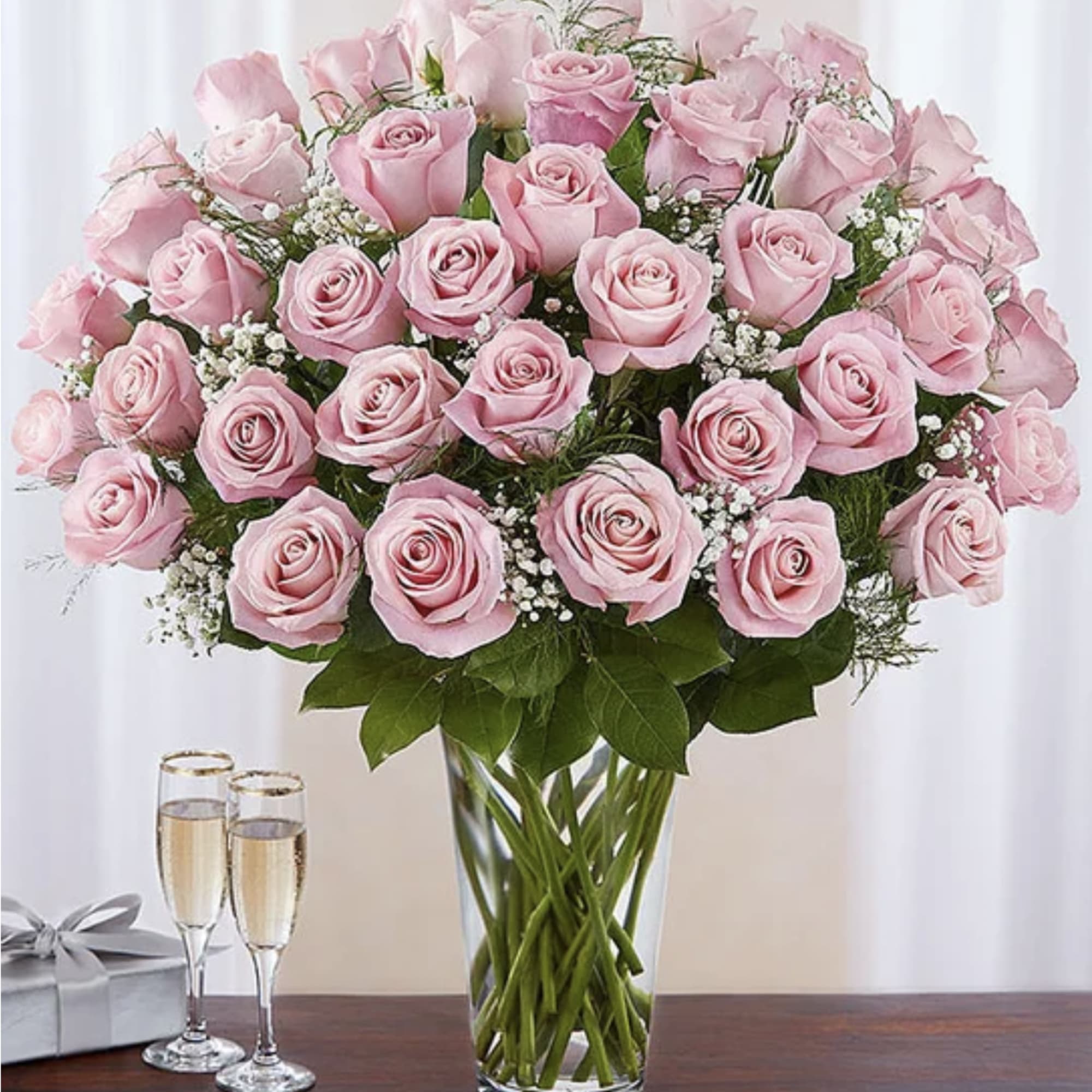 Large bouquet of pink roses in a clear glass vase beside two champagne flutes and a gift box
