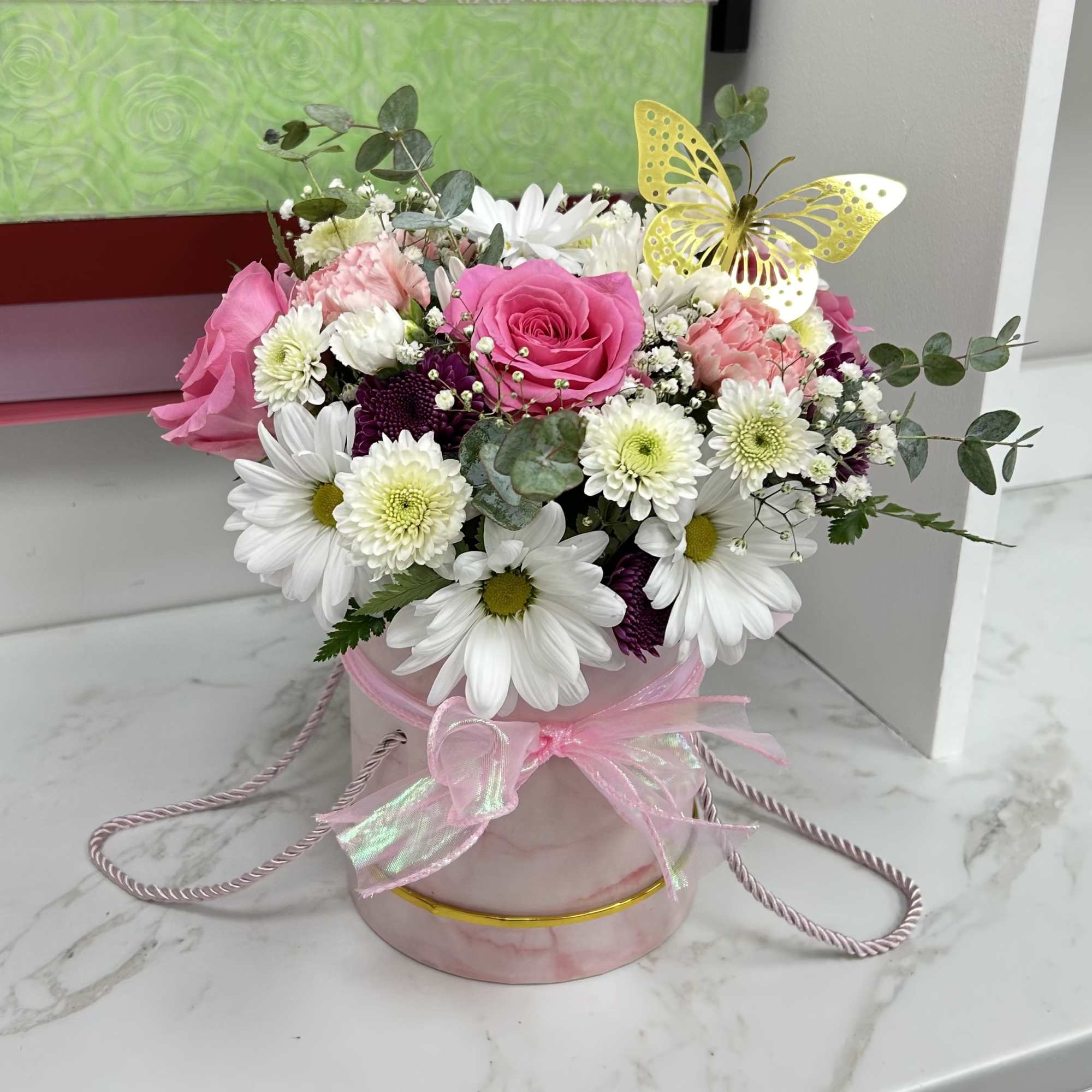 Pink and white flower arrangement with daisies and roses in a pink hatbox with a yellow butterfly pick