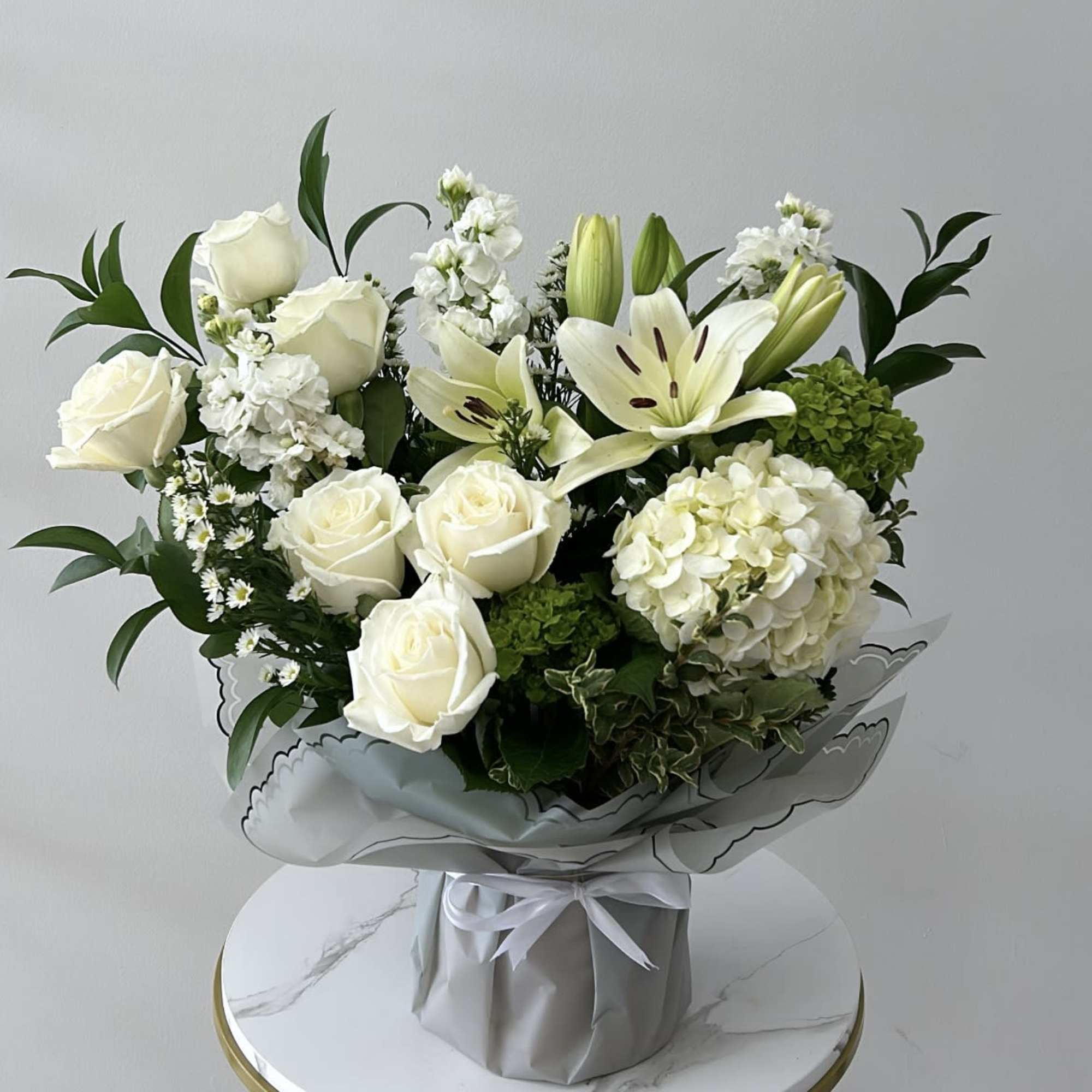 White roses and lilies arranged in a wrapped bouquet with green accents