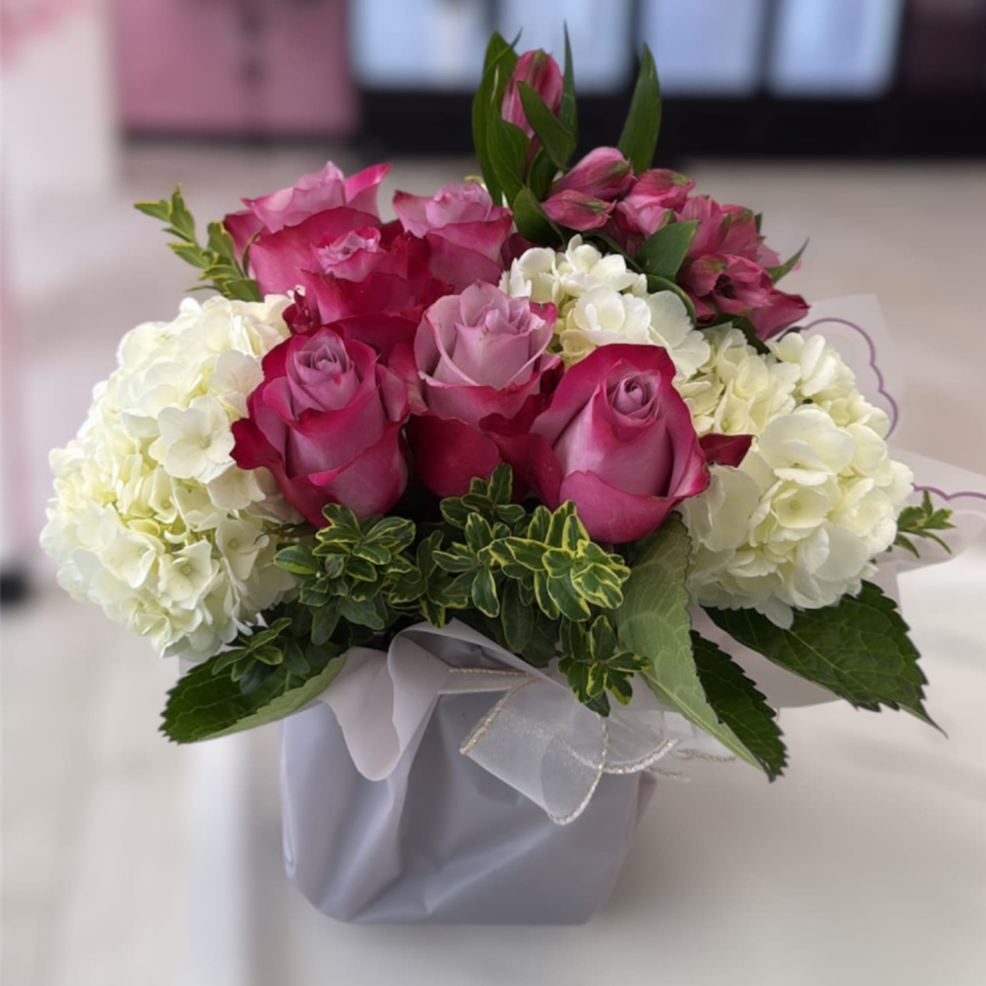 Compact arrangement of pink and lavender roses with white hydrangeas in a square vase with ribbon.
