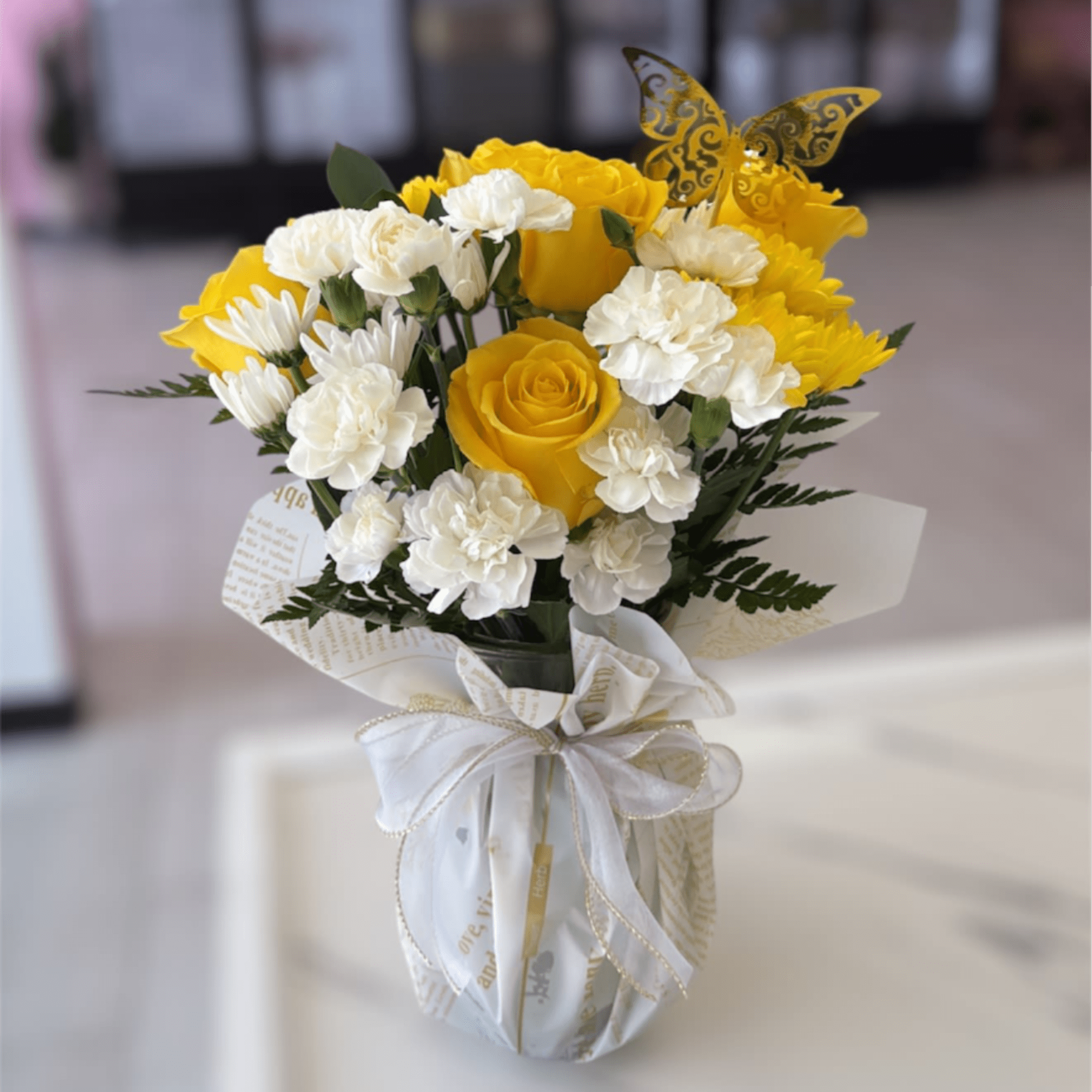 Yellow roses and white carnations in a wrapped vase with a butterfly pick