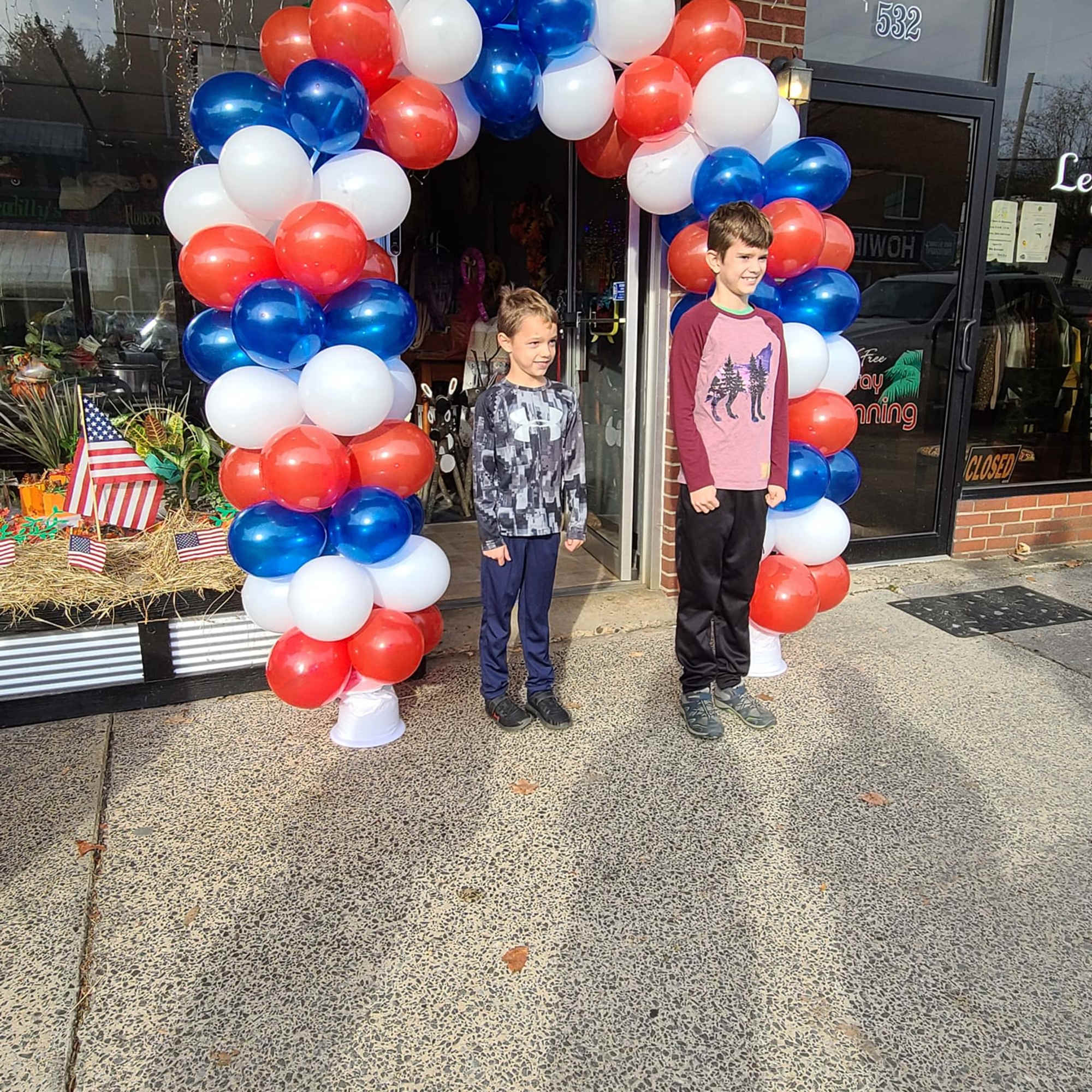 Red, white, and blue balloon arch outside a storefront with two boys standing underneath.