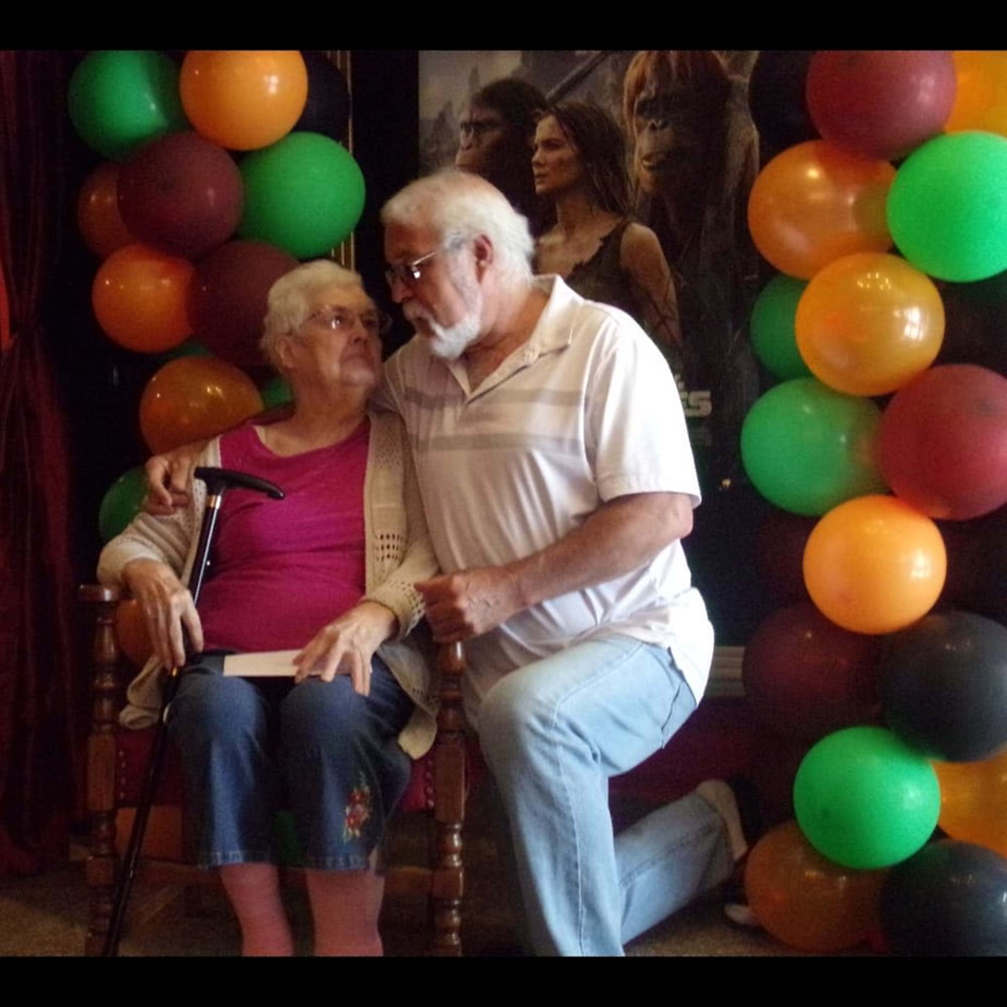Two elderly people seated between colorful balloon clusters