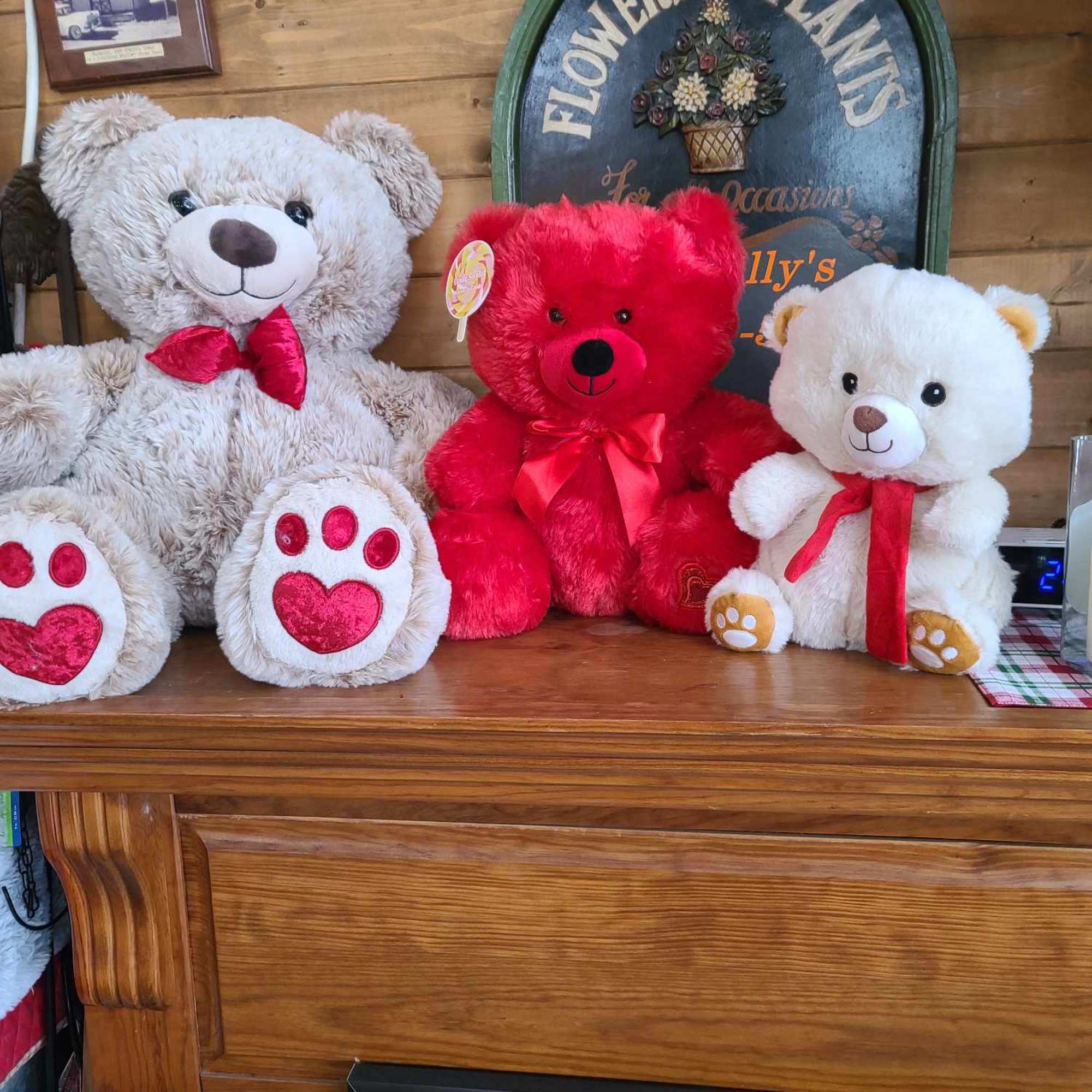 Three teddy bears with red ribbons sit on a wooden mantel.