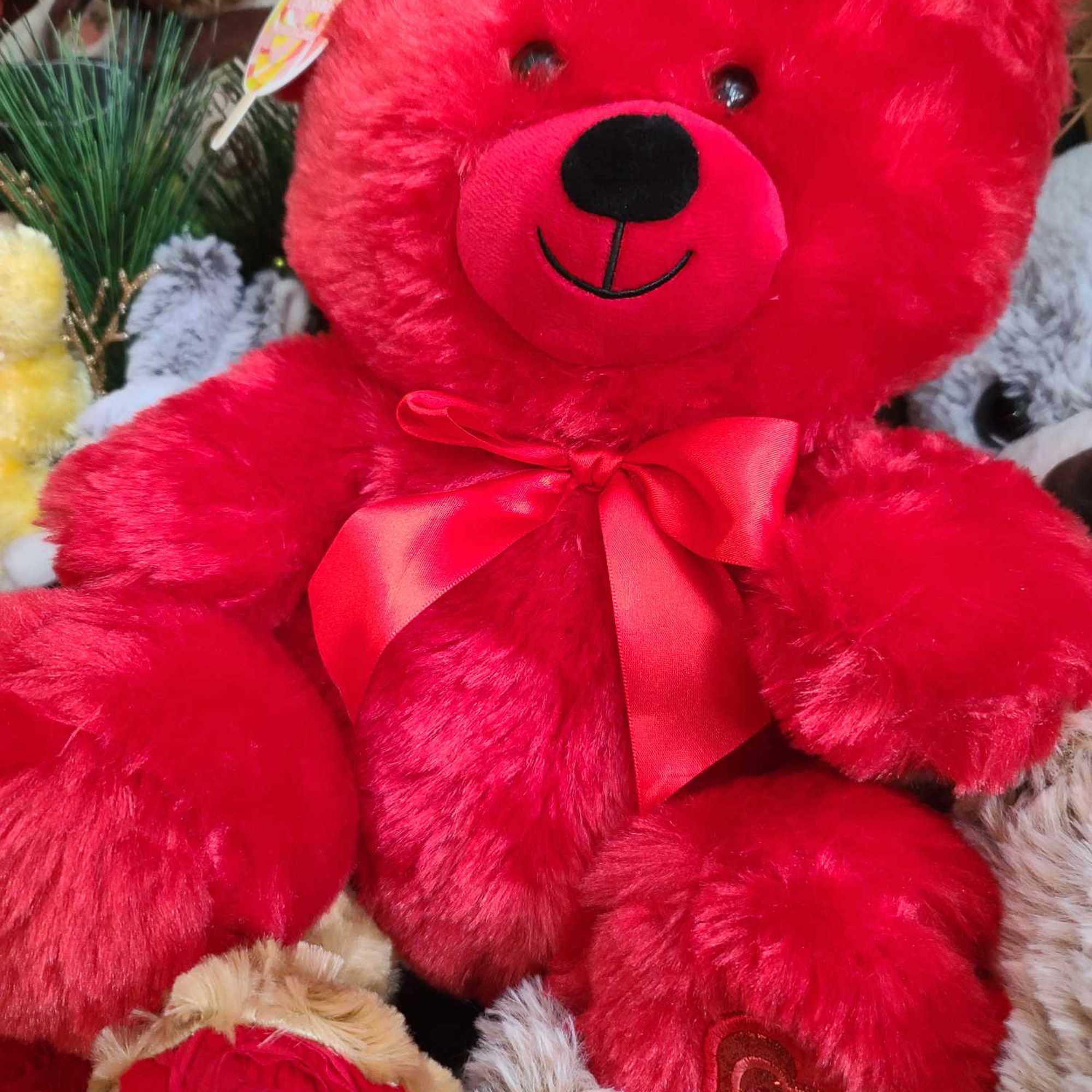 Bright red teddy bear with a satin bow, surrounded by plush toys and roses