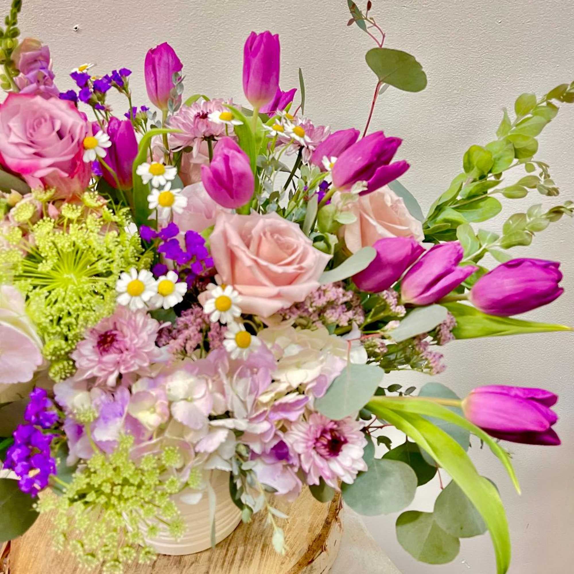 Mixed bouquet of pink roses, purple tulips, and small daisies in a white container