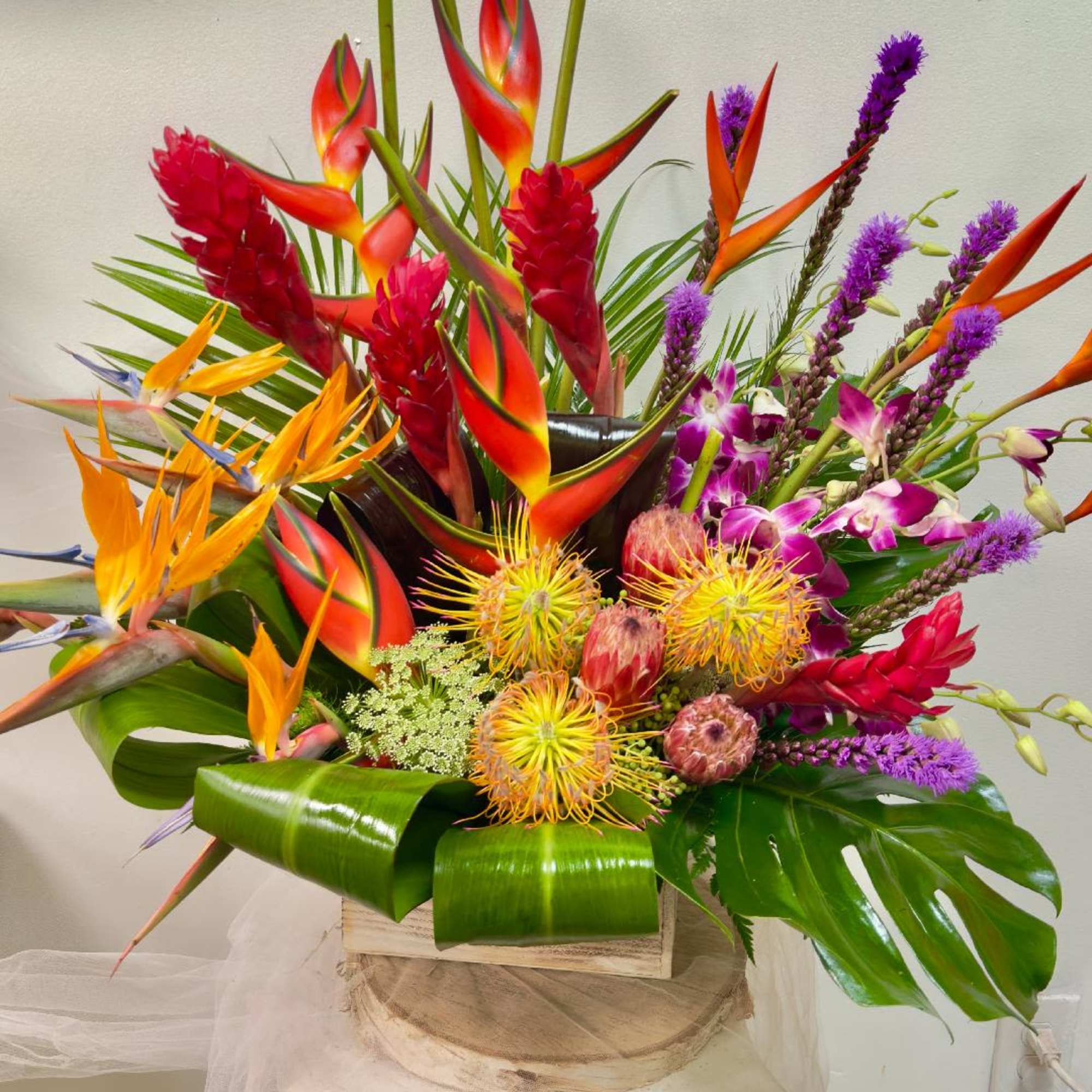 Tropical arrangement with orange heliconia, red ginger, and purple orchids in a wooden box